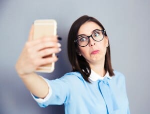 Funny businesswoman making selfie photo on smartphone. Wearing in blue shirt and glasses. Standing over gray background